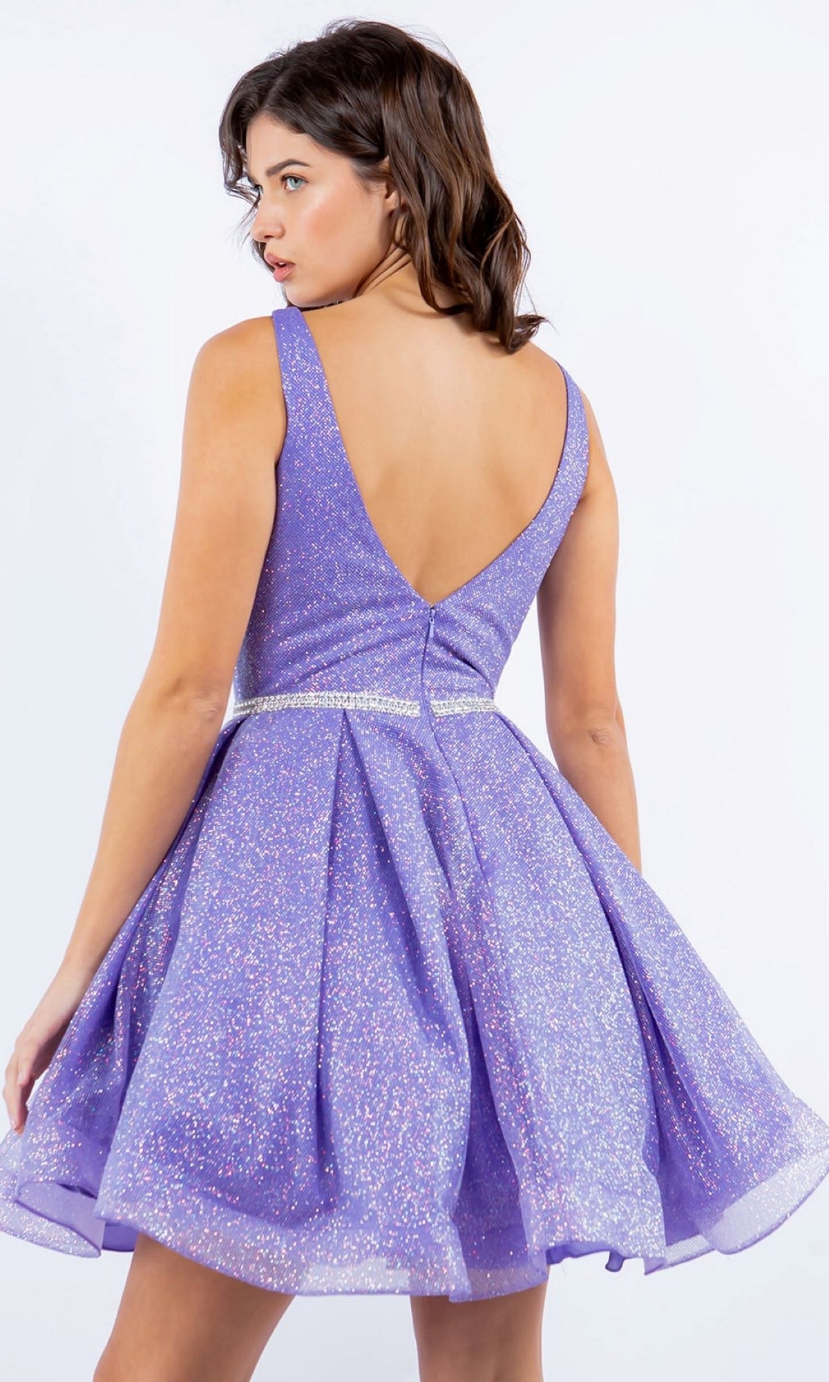 A woman stands facing away against a plain light background, wearing the Short Glitter Princess Homecoming Dress 8047J—a sleeveless, sparkly lavender dress with a V-back and flared skirt.